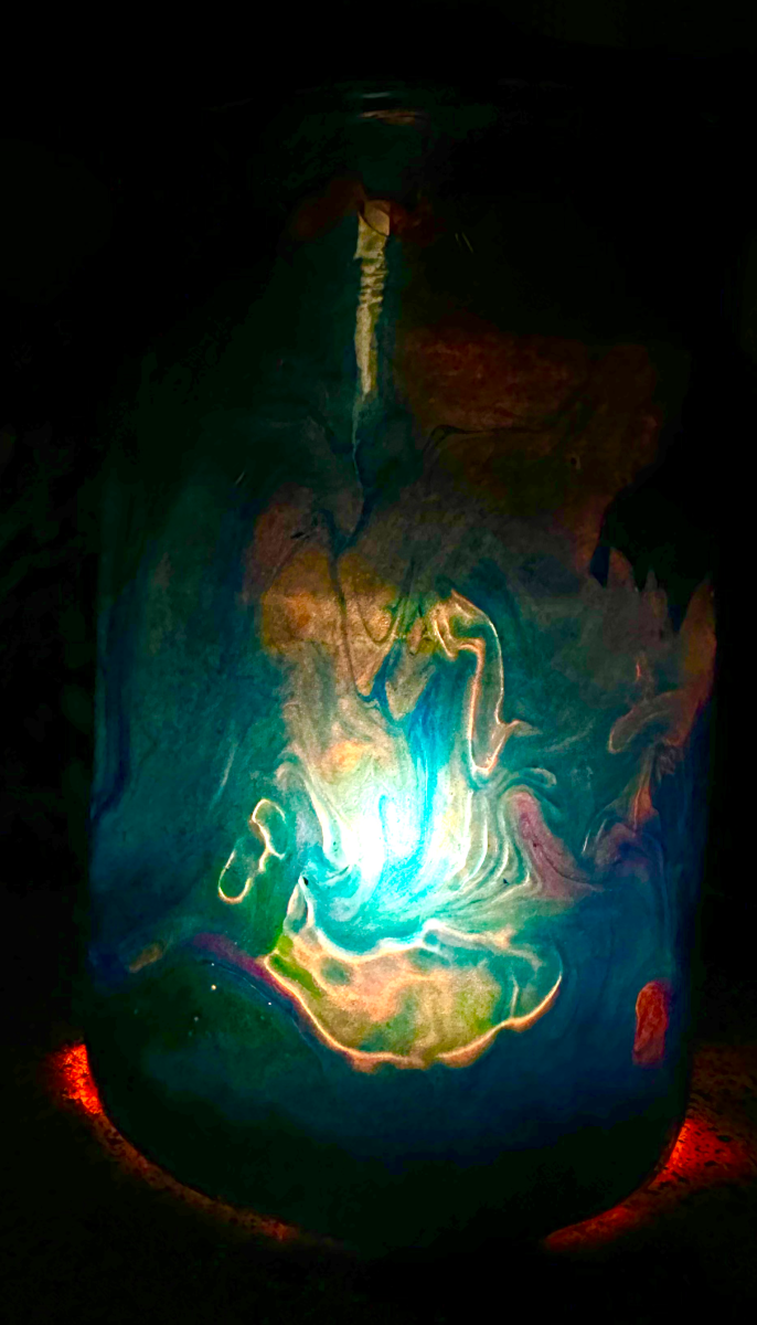 The candle jar lit from within in a dark room — teal and amber glow through the skin